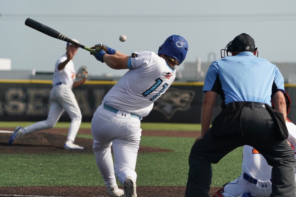A baseball player in a blue uniform swings at a pitched ball during a game, while an umpire and other players look on.