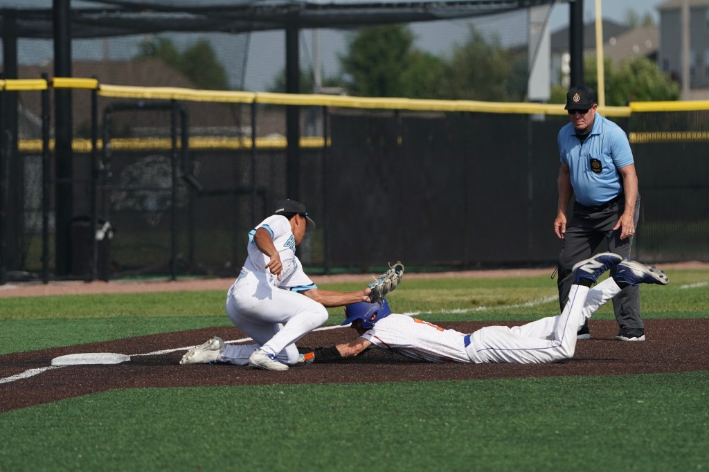 A baseball player in white slides into base while being tagged by a defender in blue. An umpire watches the play.
