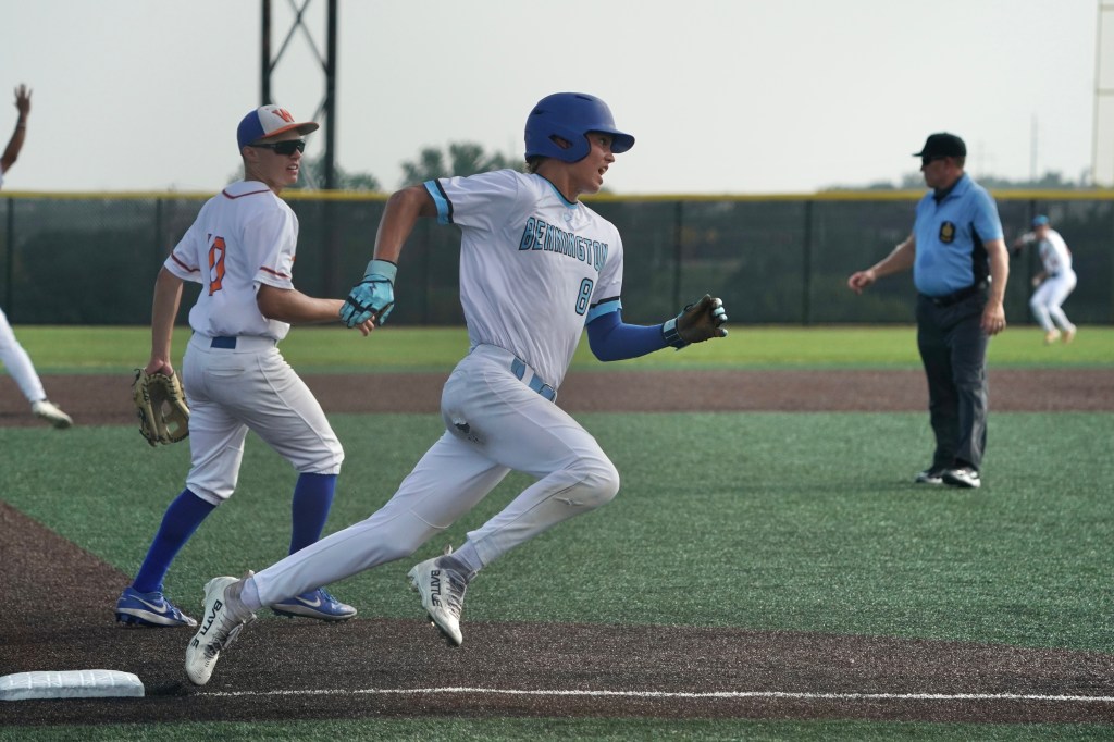 A young baseball player in a blue uniform sprinting towards first base during a game, with an opposing player in a white uniform in the background and an umpire observing the play.