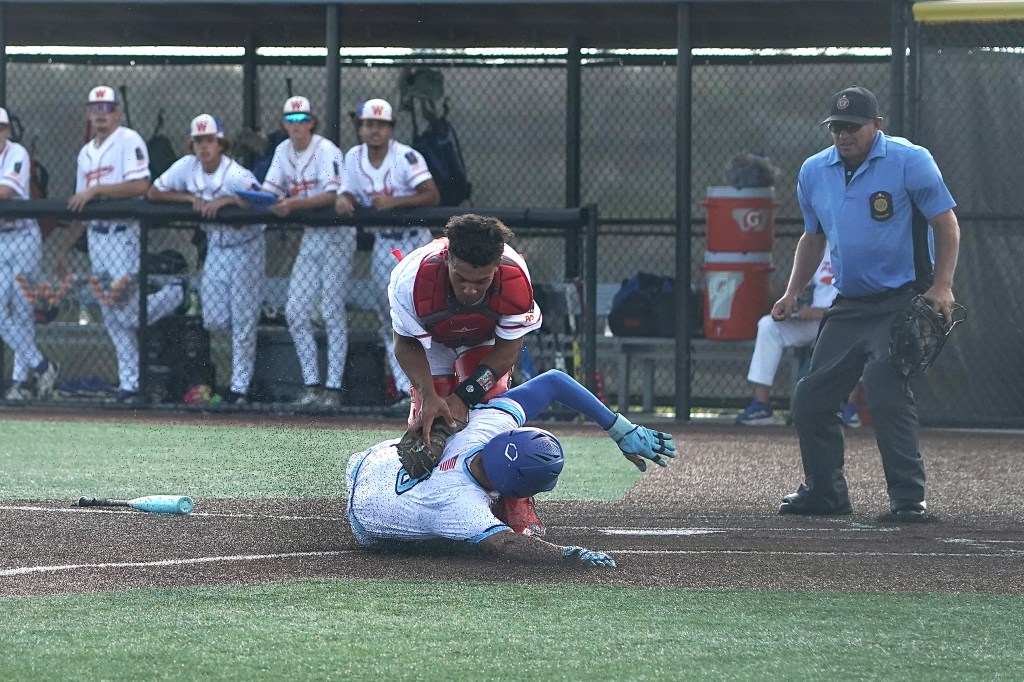 A baseball player in a blue uniform is sliding into home plate while a catcher in red prepares to tag him. An umpire watches the play closely, with players in uniforms visible in the background.