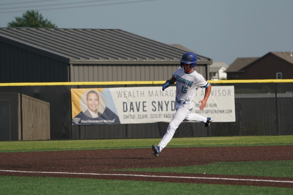 A young baseball player running towards home plate, wearing a blue helmet and a white uniform with teal accents, on a sunny day with a sports field background and a banner advertising financial services in the distance.