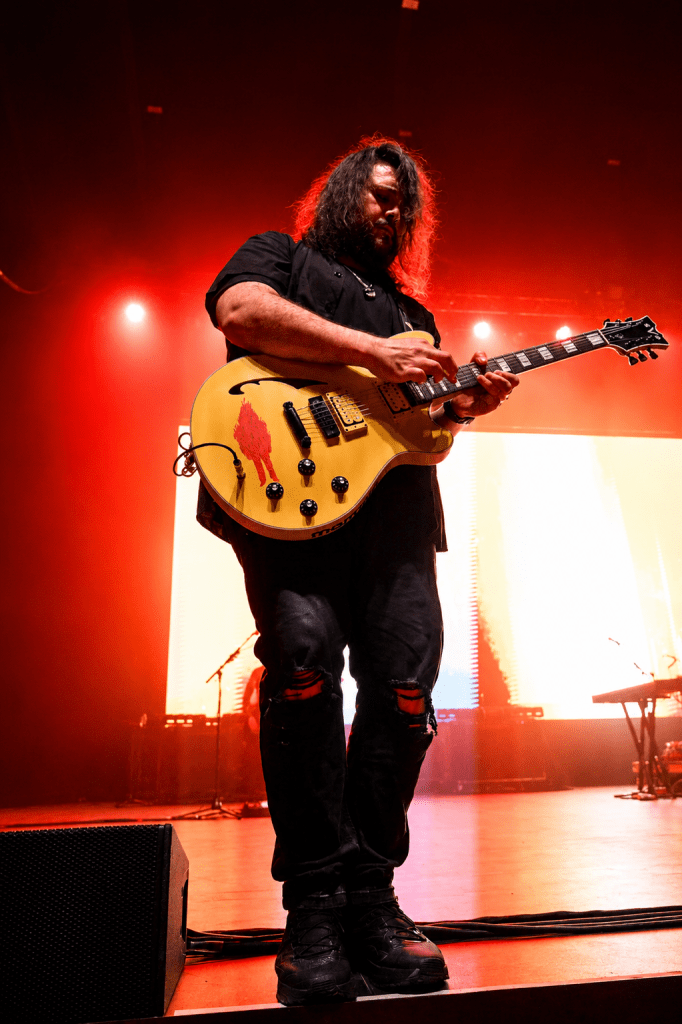 Musician playing a yellow electric guitar on stage with red lighting.