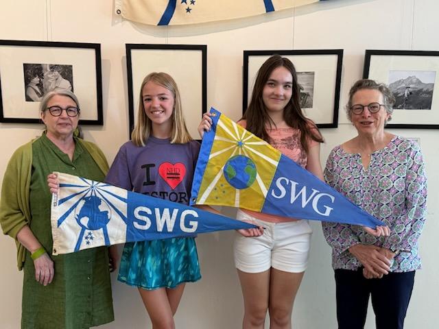 Four women standing indoors, two holding colorful flags with the letters 'SWG' and a globe design. The background features black and white photographs.