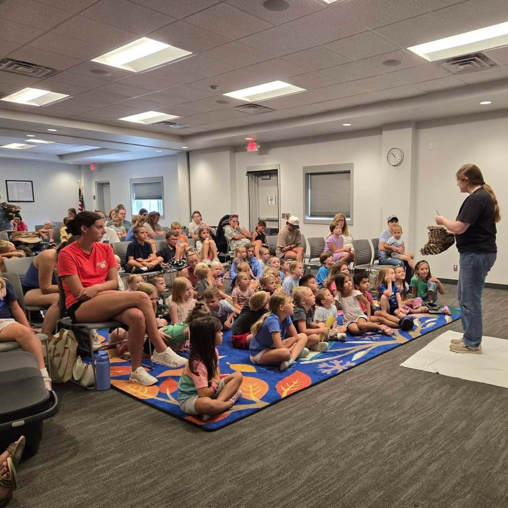 A presentation in a community room with a large group of children sitting on the floor, engaged and listening to a speaker who is holding a basket. The room features chairs and a colorful rug.