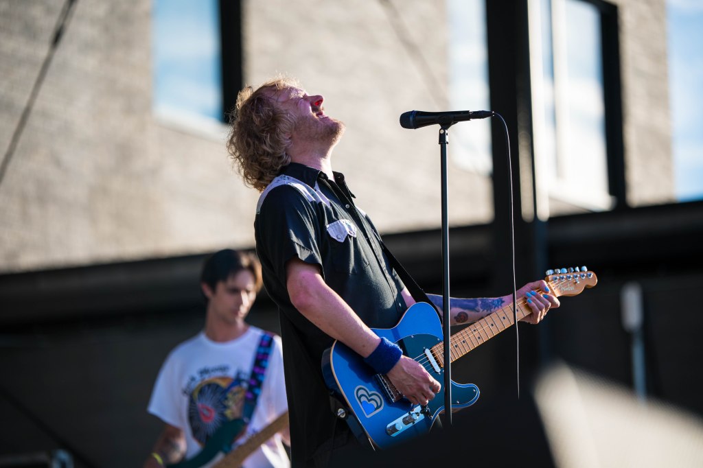 A singer performing on stage with a blue electric guitar, passionately singing while a guitarist plays in the background.