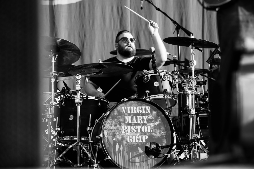 A drummer performing on stage, playing a black drum set with the band name 'Virgin Mary Pistol Grip' on the bass drum, captured in black and white.