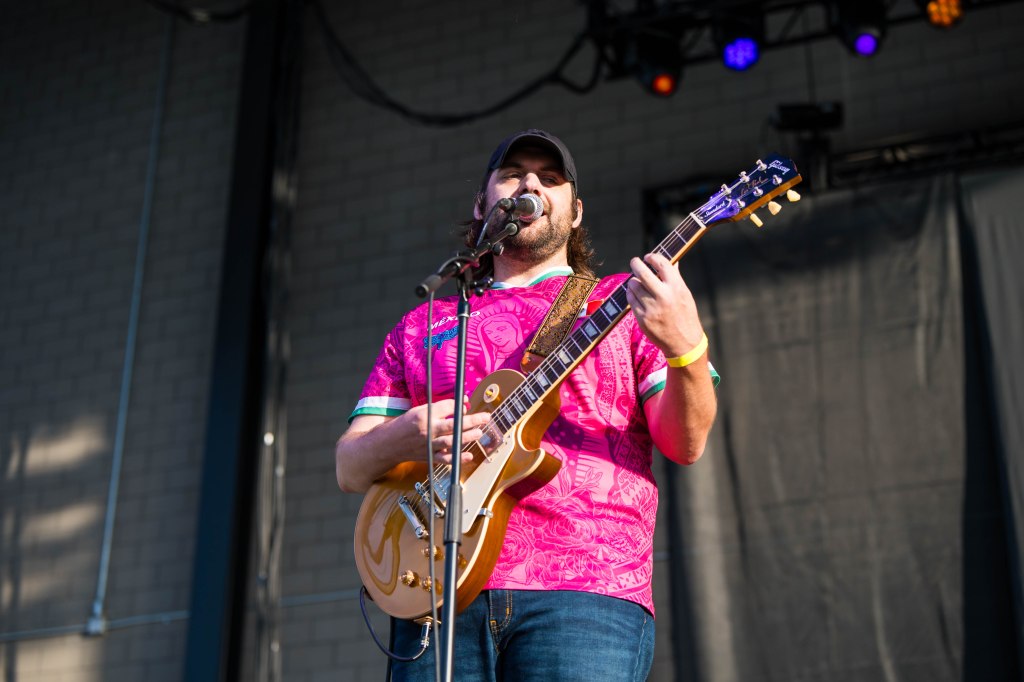 Musician performing on stage, wearing a colorful pink shirt and playing an acoustic guitar.