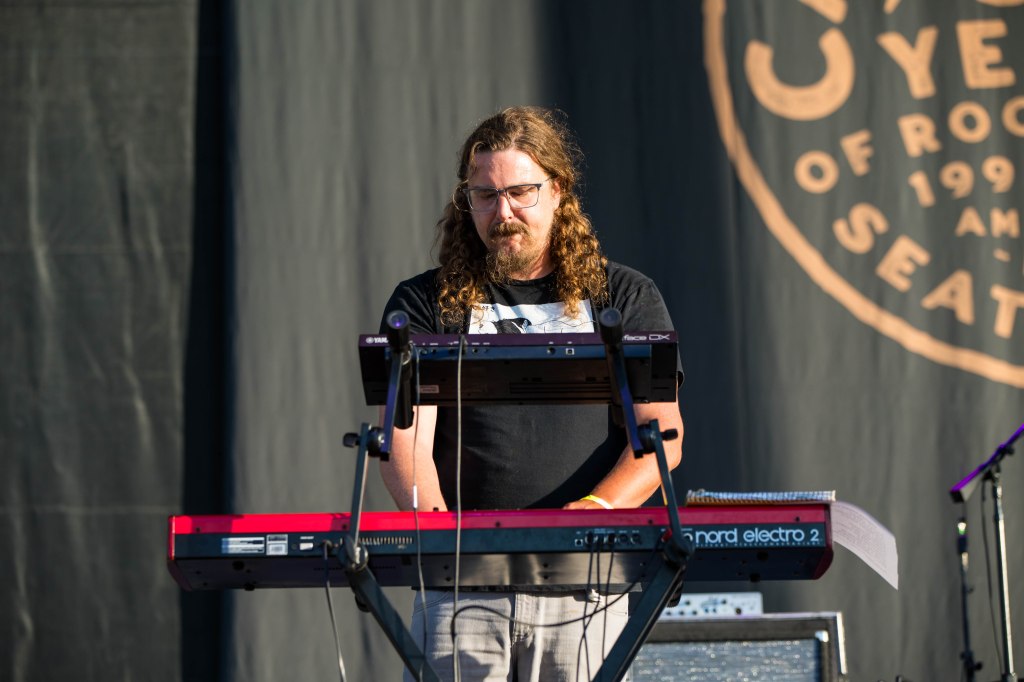 A musician playing a keyboard on stage with long curly hair and glasses, focused on the performance.