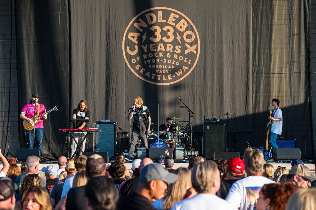 A rock band performing live on stage in front of a large audience, with a backdrop celebrating 33 years of Candlebox, a band from Seattle. The musicians include a guitarist, a keyboardist, a singer, and a drummer, all engaged in the performance.