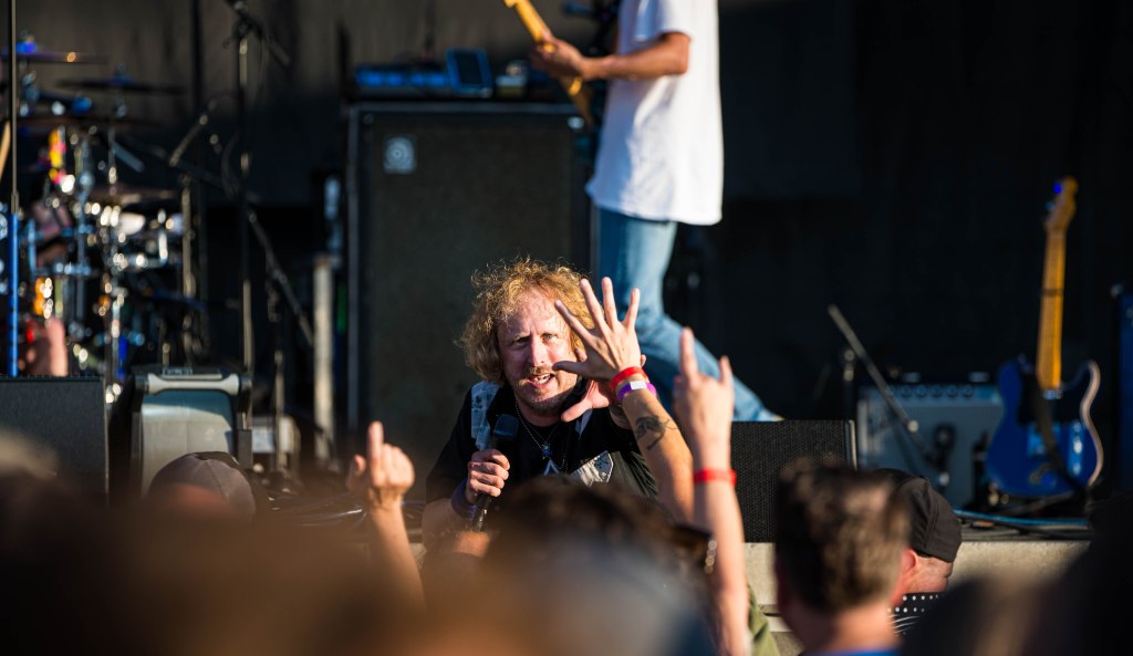 A musician speaks into a microphone while interacting with the crowd at a concert, with band members performing in the background.