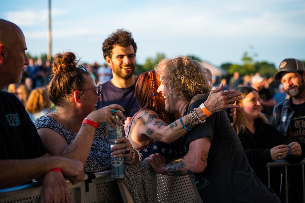 A group of people at a concert sharing joyful moments, including hugs and smiles, with a vibrant crowd in the background during sunset.
