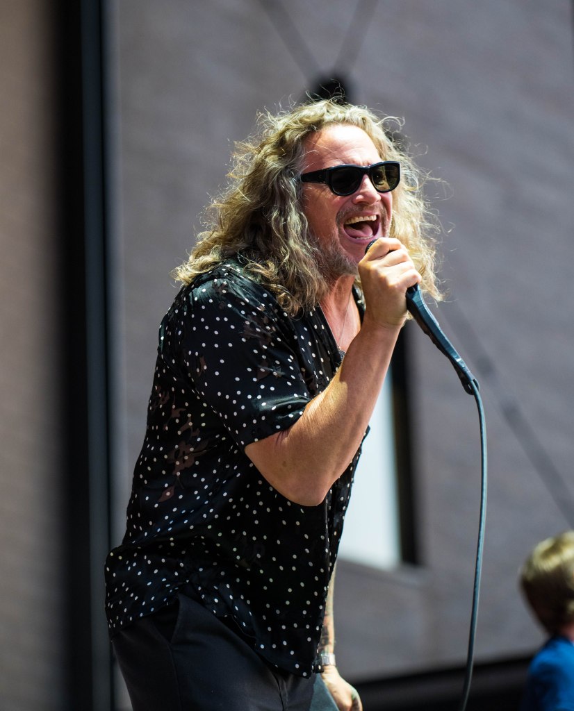 Singer performing on stage, wearing sunglasses and a polka dot shirt, passionately holding a microphone.
