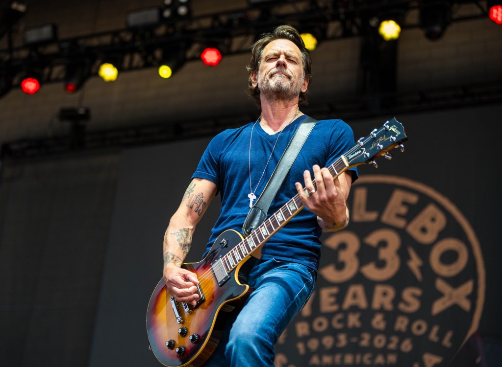 A guitarist performing on stage, passionately playing an electric guitar while wearing a blue t-shirt and jeans.