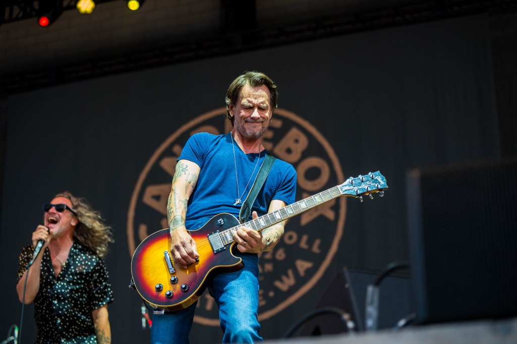 Musician playing electric guitar on stage with a singer in the background, performing at a music festival.