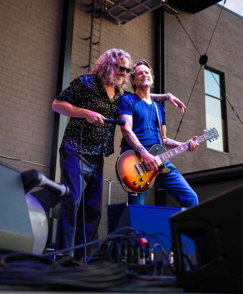 Two musicians performing on stage, one holding a microphone and the other playing a guitar, both smiling and engaging with the audience.