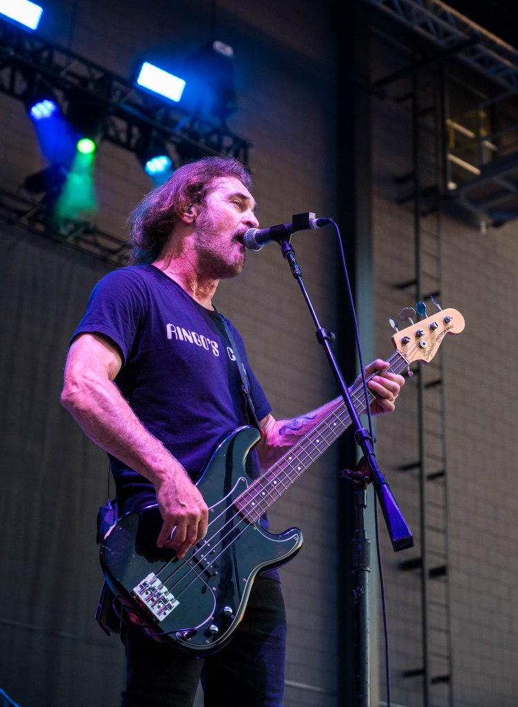 Musician performing on stage, singing into a microphone and playing a bass guitar, with stage lights in the background.