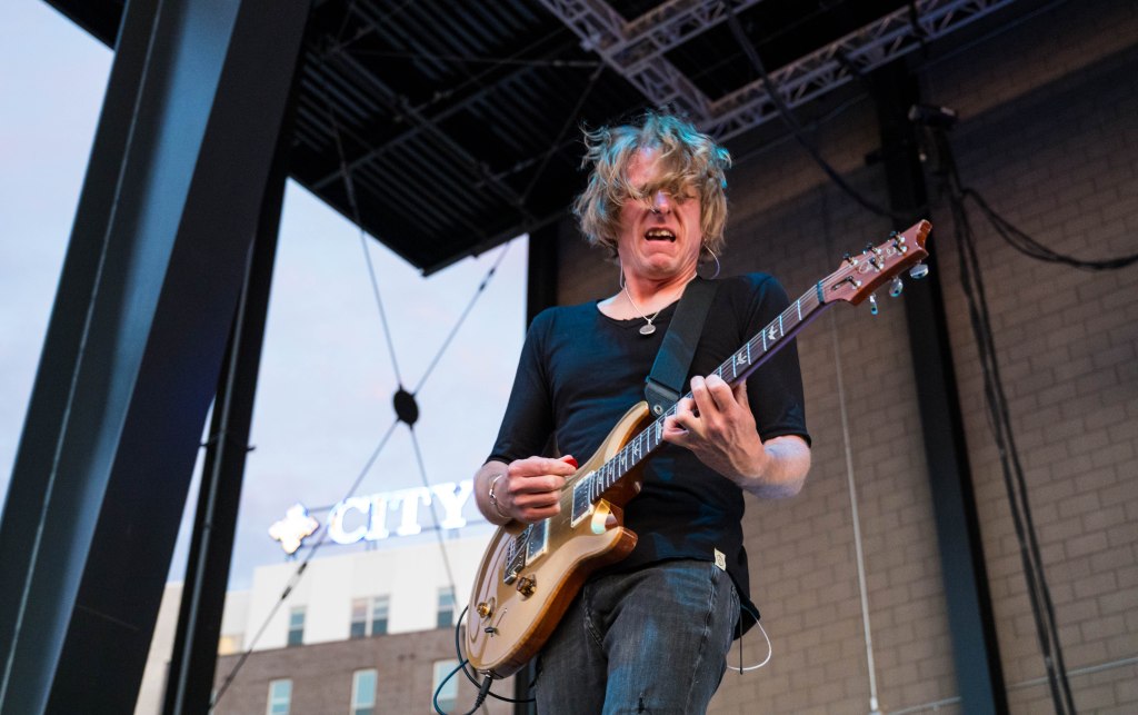 A guitarist passionately playing on stage with long hair, wearing a black shirt and jeans, against an urban backdrop with a 'CITY' sign in the background.