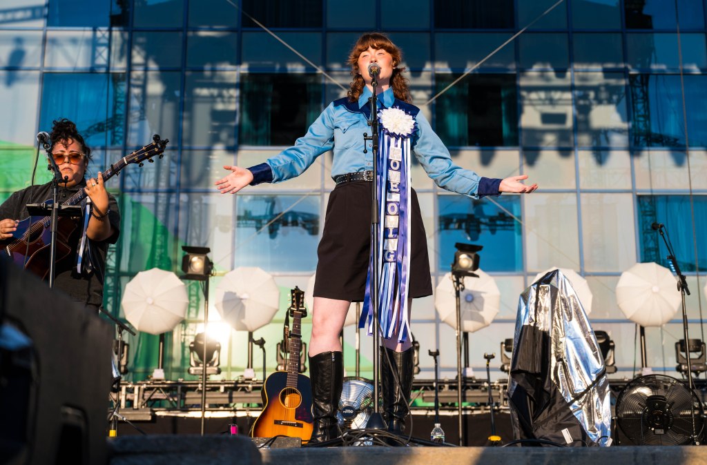 A performer on stage with arms outstretched, wearing a blue shirt and a black skirt, with a ribbon award attached. A musician with an acoustic guitar is positioned to the left, and the backdrop features large studio lights and umbrellas.