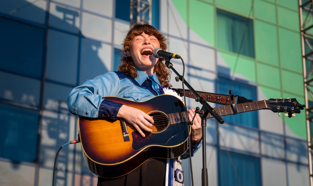 A singer-songwriter performs passionately on stage, holding an acoustic guitar and singing into a microphone, with a colorful background of windows.