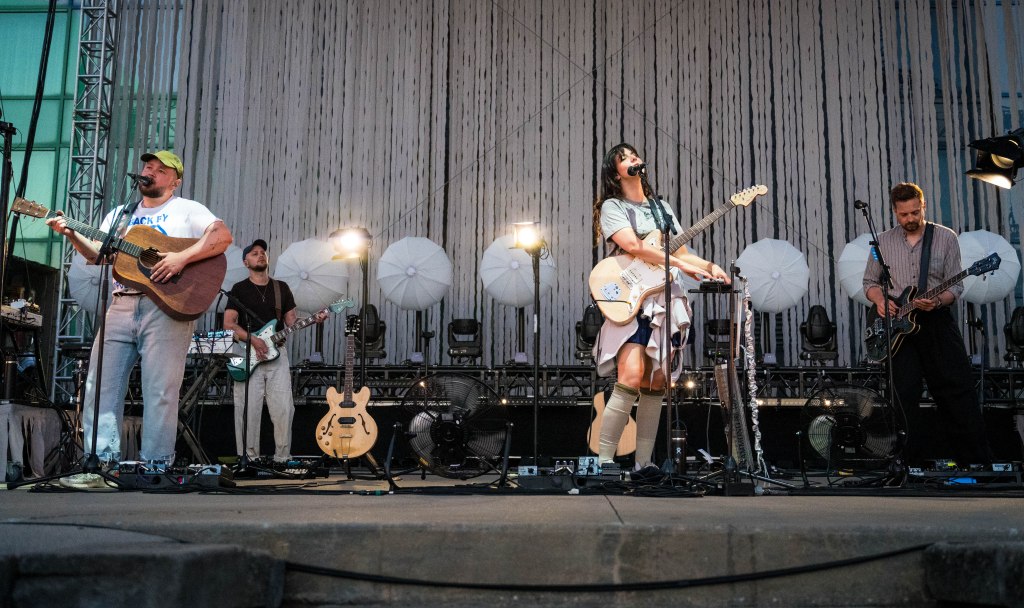 A band performing on stage, featuring four musicians: one with an acoustic guitar, one on electric guitar, a female vocalist with an electric guitar, and another on bass. The backdrop includes decorative umbrellas and stage lights.