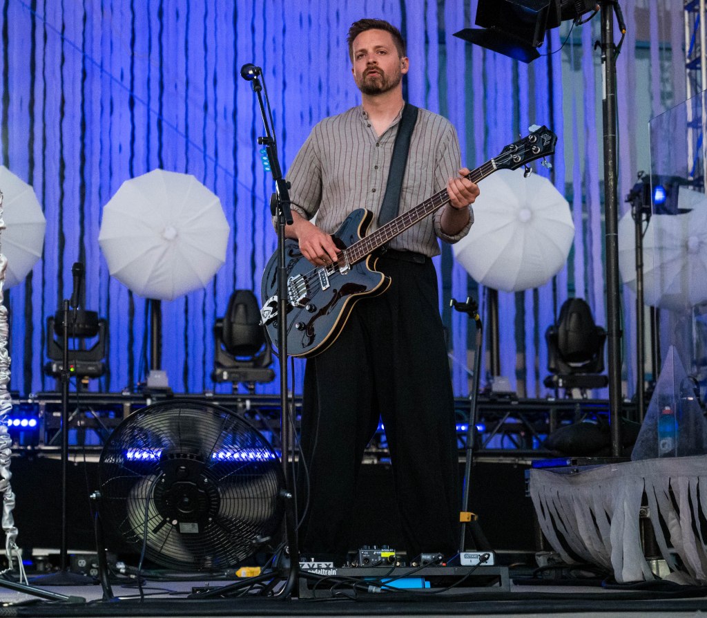 A male musician playing a bass guitar on stage, wearing a striped shirt and black pants, with a fan and stage lights in the background.