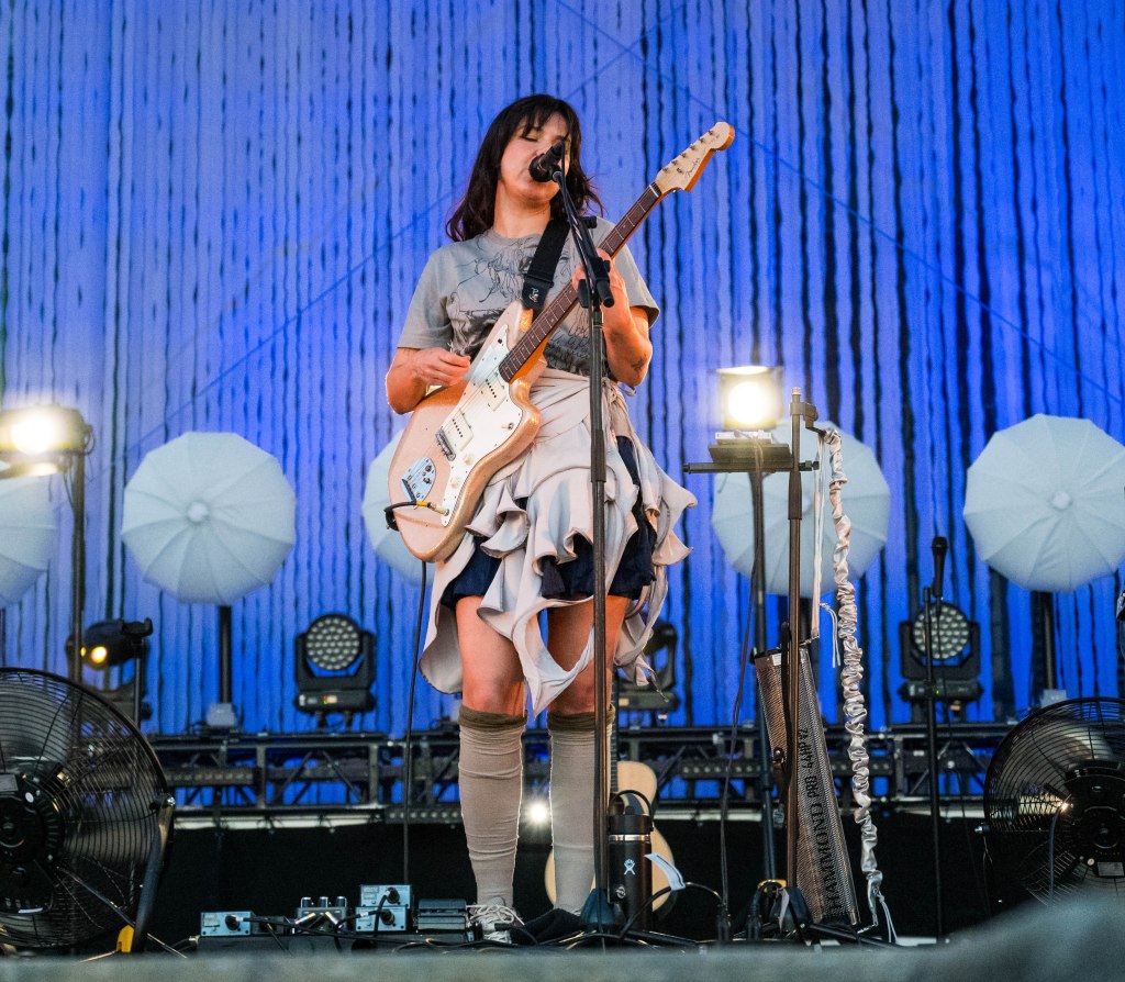 A musician performing on stage with a guitar, wearing a ruffled skirt and standing in front of a microphone, with decorative umbrellas in the background and stage lights illuminating the scene.