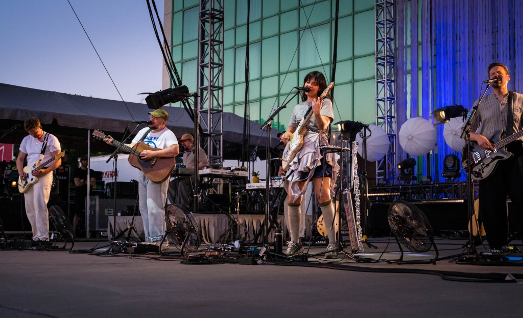 A live music performance featuring a band on stage, with four members playing various instruments, including guitars and keyboards, under stage lights during an outdoor event.