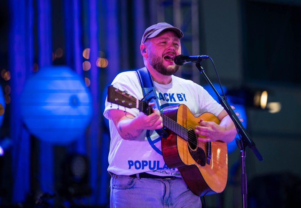 A musician performing on stage, wearing a white t-shirt with blue lettering and a cap, singing into a microphone while playing an acoustic guitar.