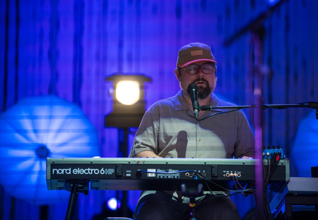 Musician playing a Nord Electro 6HP keyboard on stage, wearing a cap and glasses, with a microphone in front of him and colorful lighting in the background.