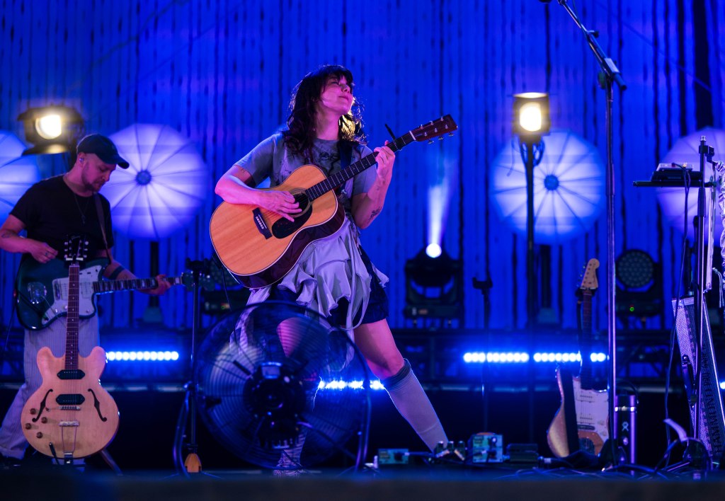 A female musician is performing on stage with an acoustic guitar, surrounded by colorful stage lights and sound equipment. A male guitarist stands nearby, partially visible.