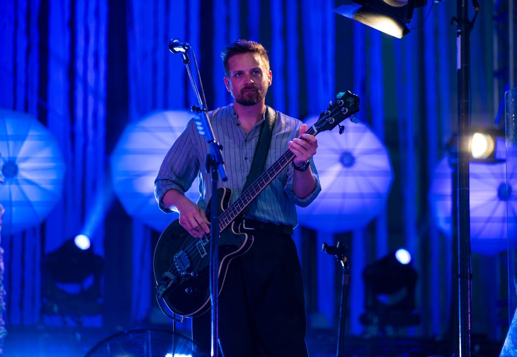 A male musician playing a black bass guitar on stage, with blue lighting and decorative lanterns in the background.