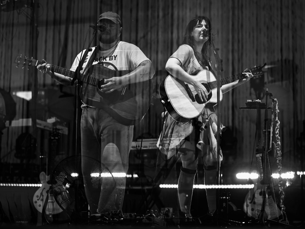 A black and white image of two musicians performing on stage. One is a man playing an acoustic guitar and singing, while the other is a woman with an acoustic guitar, also singing. They are surrounded by stage equipment and soft lighting.
