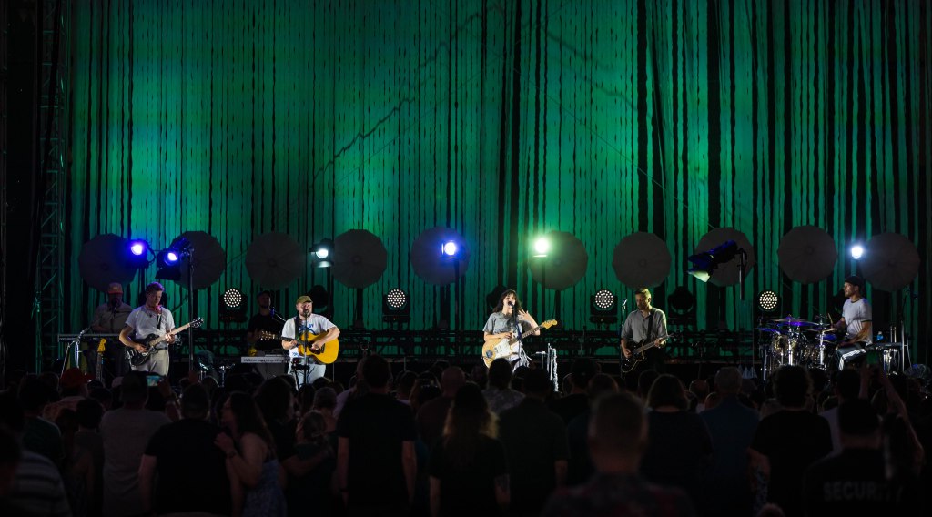 A band performing on stage with a vibrant green backdrop and illuminated spotlights, while a crowd enjoys the live music.