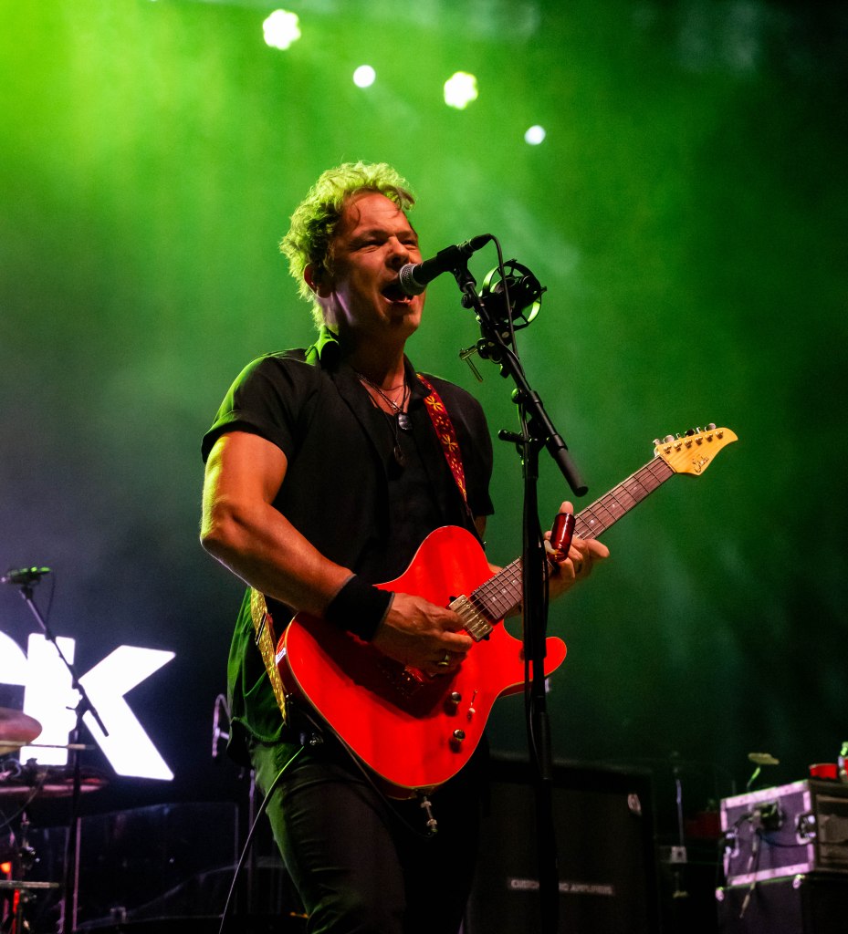 A male guitarist passionately performing on stage with a red electric guitar, microphone in front, under vibrant green stage lights.