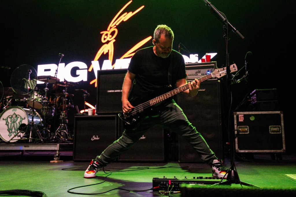 A bassist performing energetically on stage, with a large 'BIG WET' backdrop and musical equipment in the background.