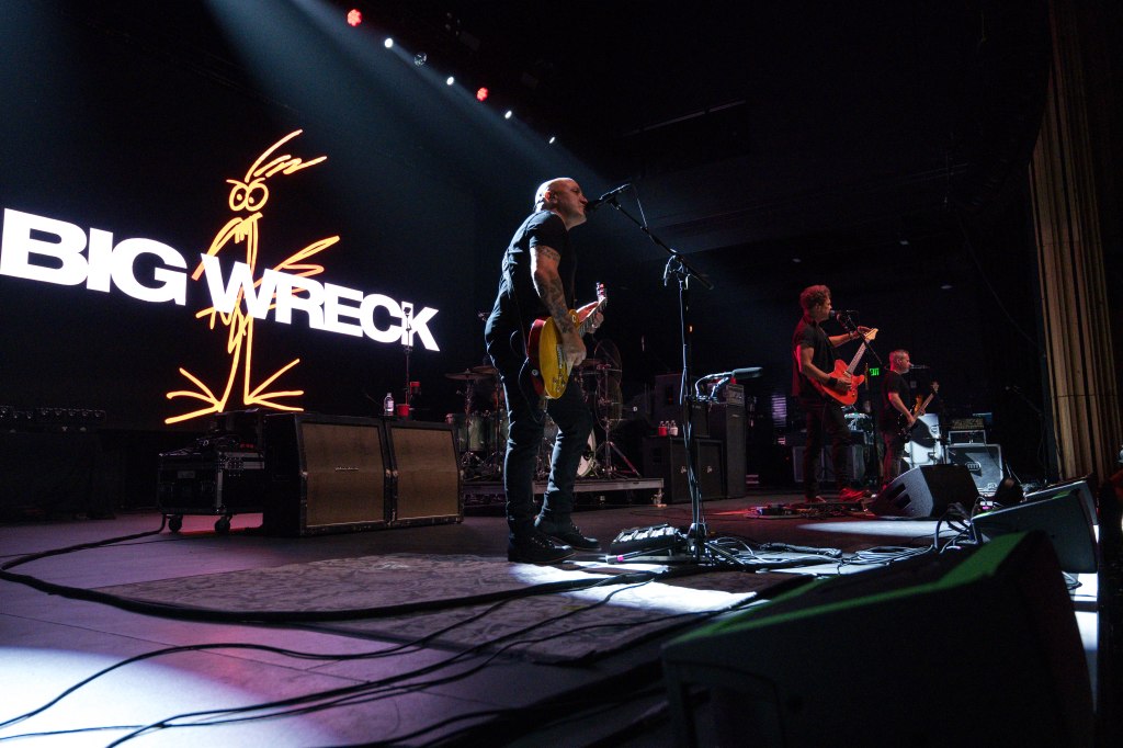 A live band performance featuring four musicians on stage, with a large backdrop displaying the words 'BIG WRECK' and a cartoon-like drawing of a bird.