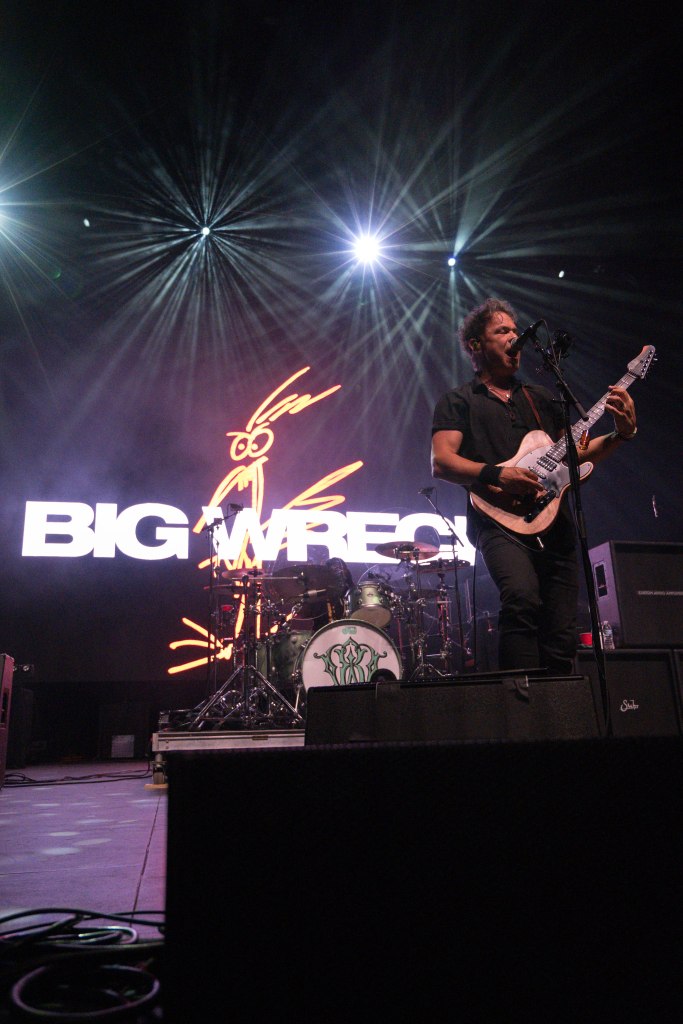 A musician performing on stage in front of a large screen displaying the text 'BIG WRECK' with a cartoon character illustration.
