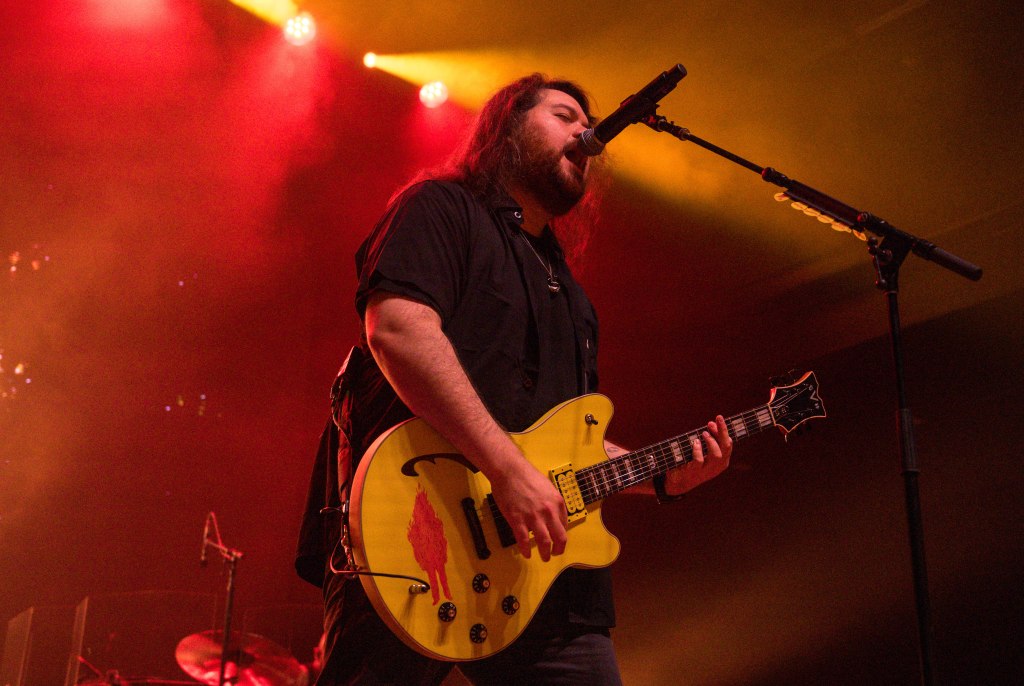 A musician performing on stage with a yellow guitar, singing into a microphone, surrounded by colorful stage lights.