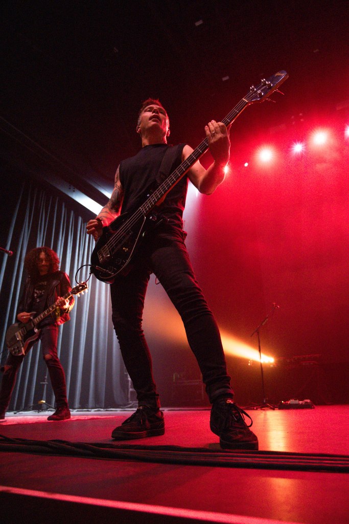 A musician performing on stage, playing a bass guitar, with dramatic lighting in shades of red and shadows. Another band member is visible in the background.
