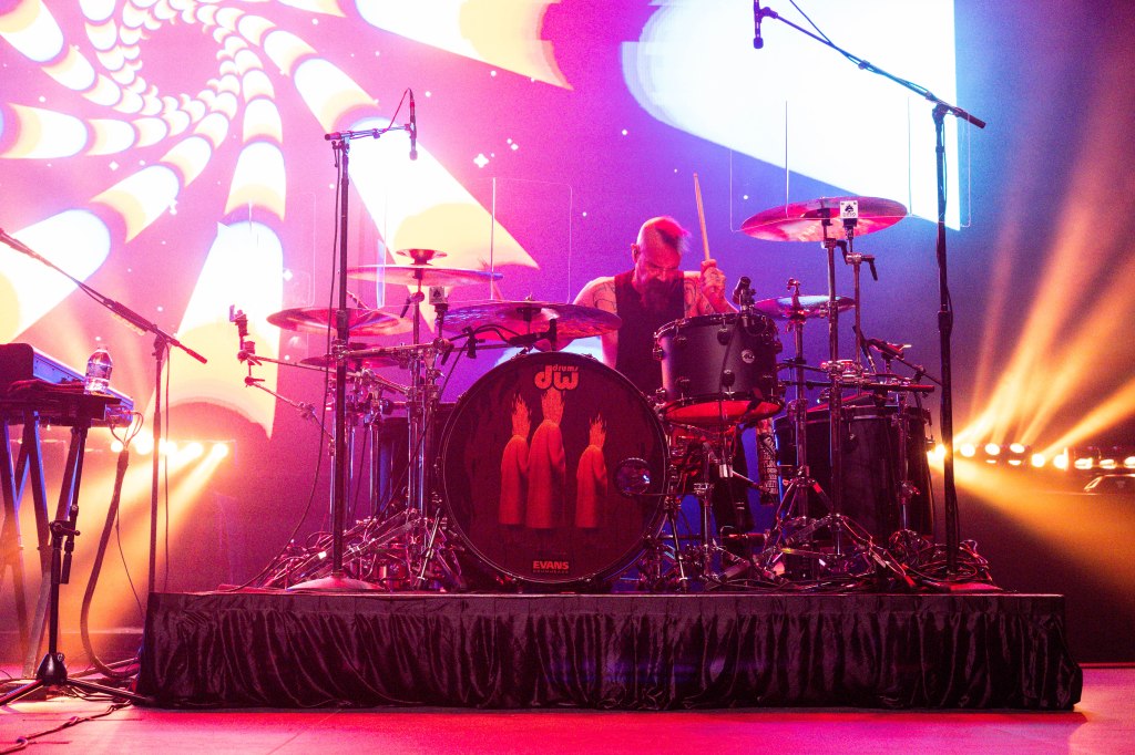Drummer performing on stage with colorful psychedelic background lights.