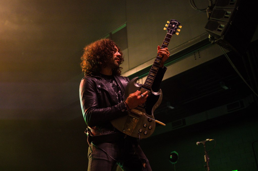 A musician performing on stage, playing a guitar. The artist has curly hair, wears a leather jacket, and stands under stage lights, with a focus on their passionate expression and dynamic pose.
