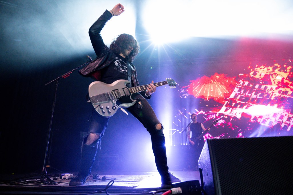 A guitarist performing live on stage, raising one hand while playing an electric guitar, with colorful lights and a band member in the background.