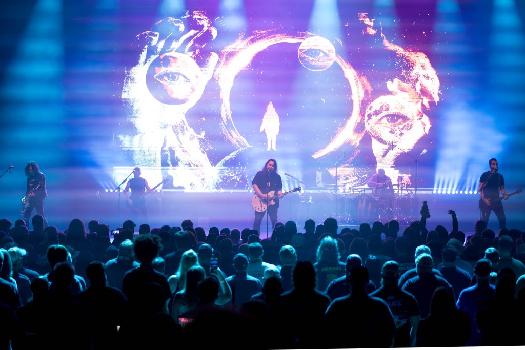 A musical performance on stage with a band playing in front of a large audience. The background features vibrant visual effects, including abstract images and colorful lighting.