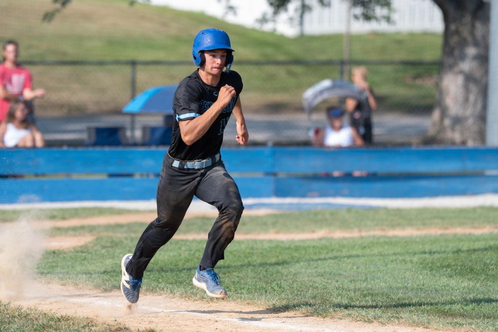 A baseball player wearing a blue helmet and black uniform runs towards home plate on a dusty field, with spectators in the background.