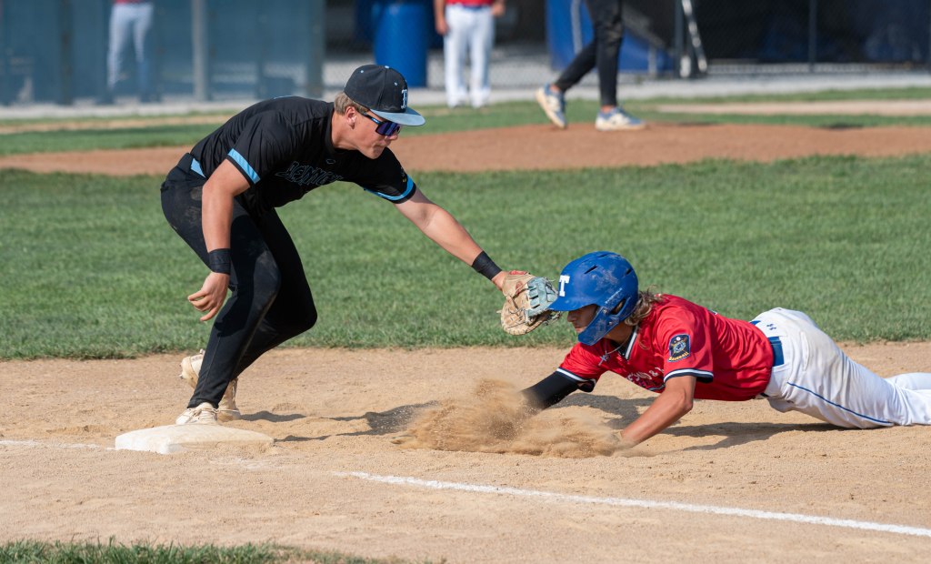 A baseball player in a black uniform attempts to tag another player in a red uniform who is sliding into first base on a sandy field.