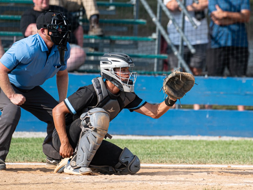 A baseball catcher in protective gear crouches behind home plate, wearing a helmet, while an umpire in a blue shirt stands ready to make a call.