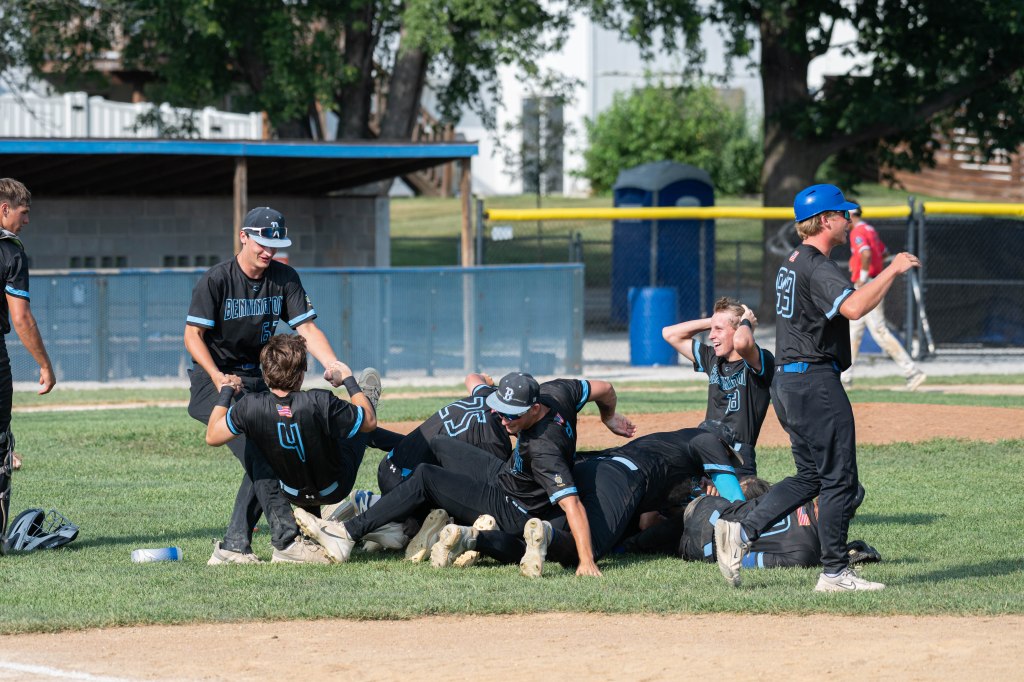 A group of young baseball players in black uniforms celebrating on the field, some piling on top of each other in joy after a win.