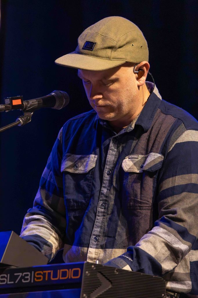 A musician playing a keyboard on stage, wearing a cap and a plaid shirt, focused on the performance.