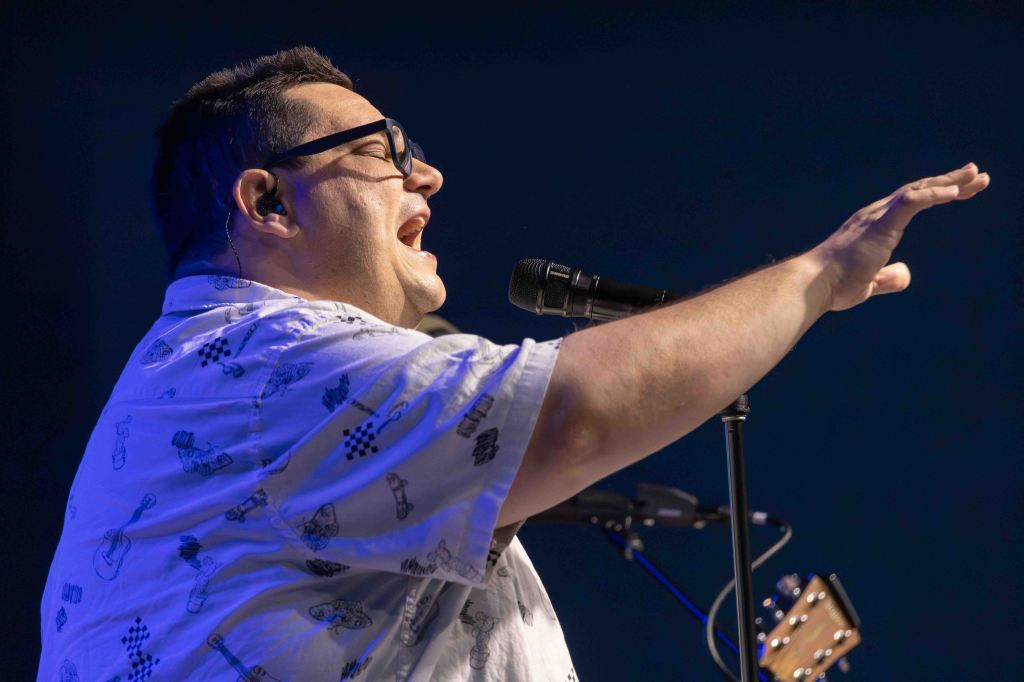 A singer with glasses passionately performing on stage, gesturing with one hand while holding a microphone. He is wearing a patterned shirt and there is an acoustic guitar in the background.