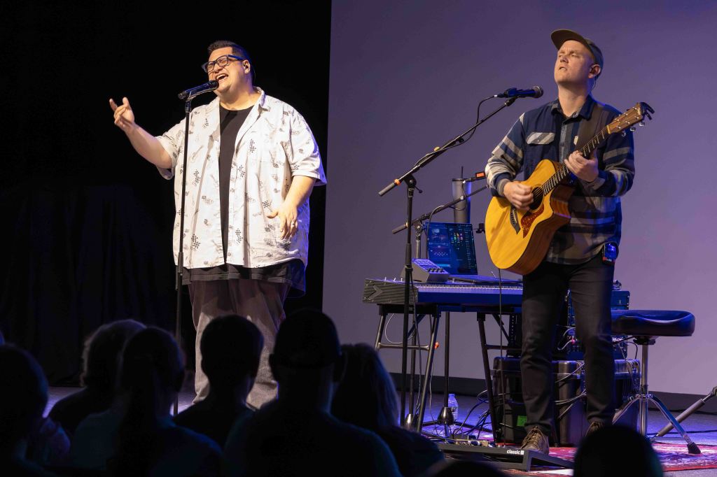 A live music performance featuring a singer with glasses and a patterned shirt, passionately singing into a microphone, alongside a guitarist playing an acoustic guitar.